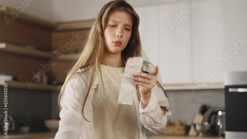 Sad young woman looking at check next to table with fresh vegetables from supermarket and compares prices
at home kitchen Upset female have not money after expensive food shopping indoors
