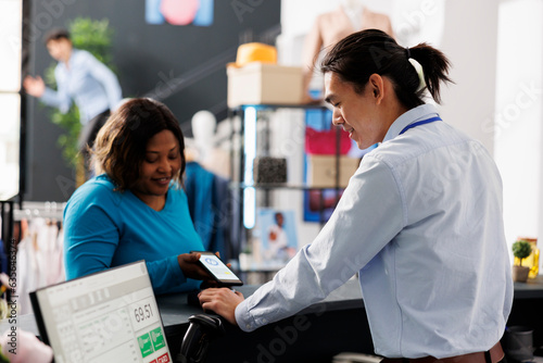 African american customer putting phone on pos terminal paying for stylish clothes in boutique after discussing items fabric with employee. Woman standing at retail store counter buying casual wear photo