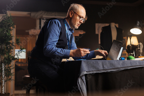 Fashion designer buying sewing materials online for upcoming customer comission tailoring modifications. Elderly couturier imputing credit card purchasing details on laptop in atelier studio