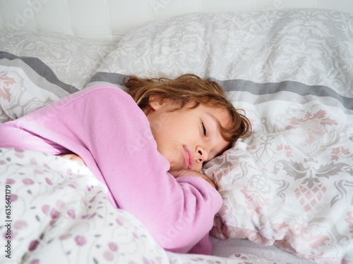 A beautiful caucasian girl of 8 years old with blond hair, dressed in pink pajamas, sleeps on a bed with a fluffy blanket, hugging a pillow. Soft light of the morning sun streams through the window.