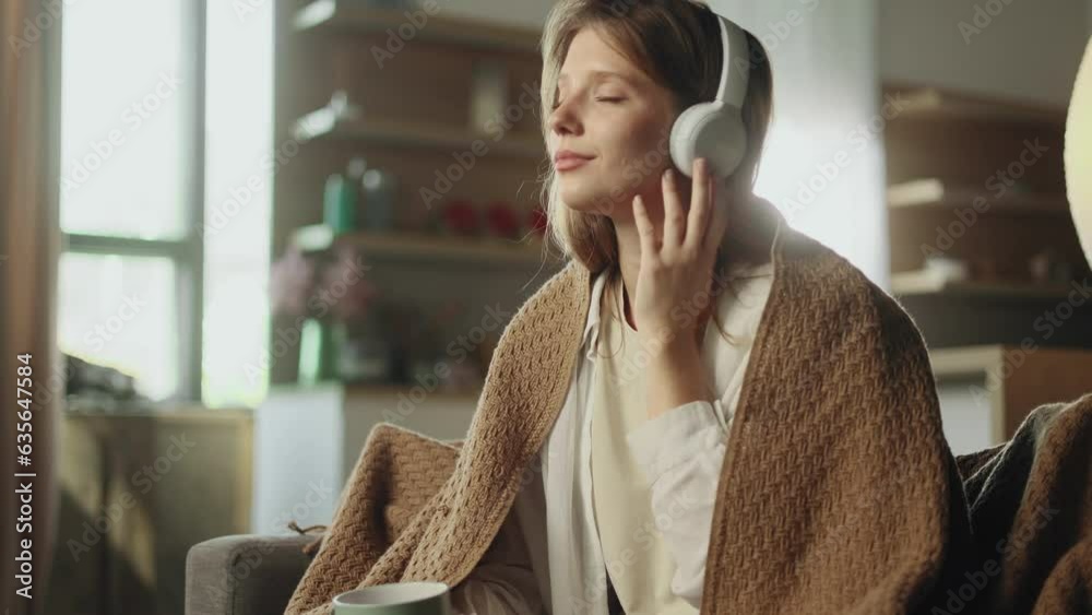 Portrait of pretty young woman under the blanket listening to music on sofa with soft light behind her indoors Smiling female with headphones hold cup of hot drink enjoying leisure time at home alone