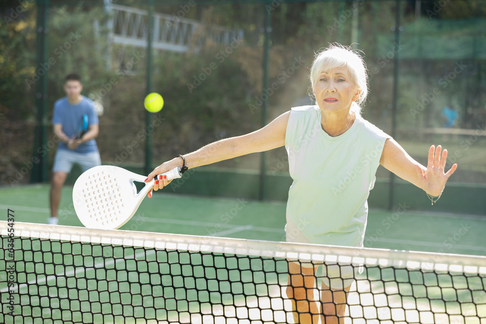 Concentrated elderly woman padel player hitting ball with a racket on a hard court