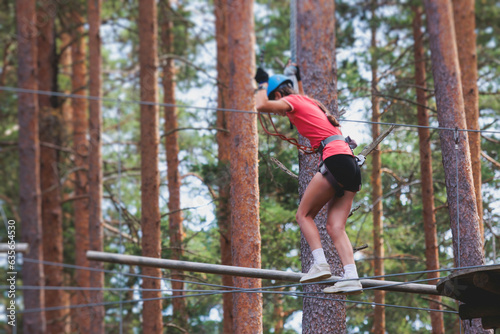 View of high ropes course, process of climbing in amusement acitivity rope park, passing obstacles and zip line on heights in climbing safety equipment gear between the trees om heights, summer day