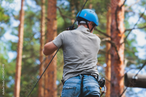 View of high ropes course, process of climbing in amusement acitivity rope park, passing obstacles and zip line on heights in climbing safety equipment gear between the trees om heights, summer day