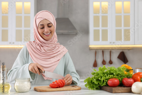 Muslim woman making delicious salad with vegetables at white table in kitchen. Space for text