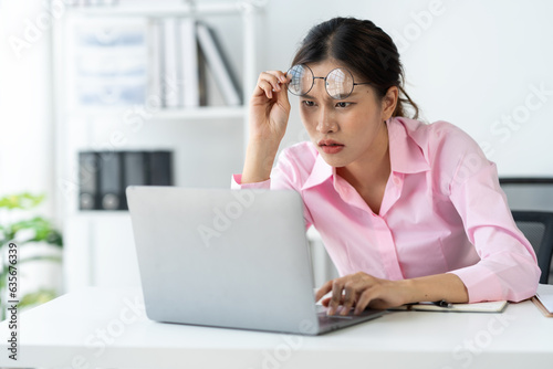 Asian businesswoman holding glasses thinking hard have pain around the eyes Tired of worrying about how to solve online document data by looking at the laptop screen in the office.