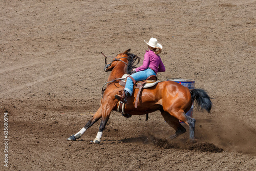 Cowgirl riding in a barrel racing completion at a rodeo. She is wearing red shirt and blue jeans and a white hat. The horse is brown. The barrel is to the right of the horse.