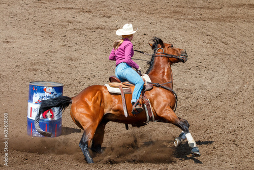 Cowgirl riding in a barrel racing completion at a rodeo. She is wearing red shirt and blue jeans and a white hat. The horse is brown. The barrel is behind the horse.