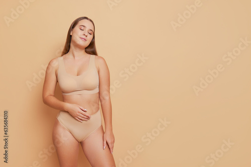 Attractive girl in beige ergonomic underwear stands in calm pose with one hand on her belly and closed eyes against studio background, natural beauty concept, copy space