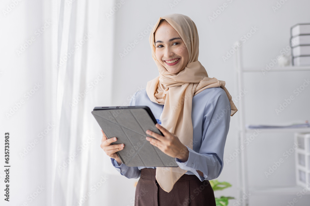 Young business Asian Business muslim woman in purple hijab using tablet and phone, standing near the window in workplace business finance concept. Young business Asian Business muslim woman in purple hijab using tablet and phone, standing near the window in workplace business finance concept.