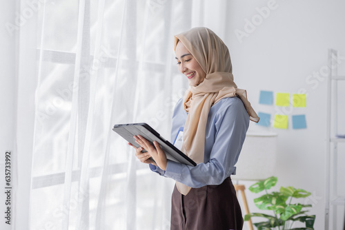 Young business Asian Business muslim woman in purple hijab using tablet and phone, standing near the window in workplace business finance concept.	