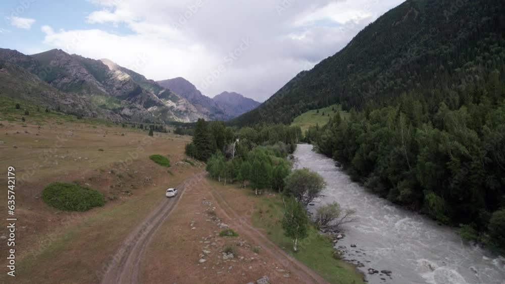 A white SUV overcomes a mountain route along a gorge along the river. Green fields with cliffs. Steep cliffs and big rocks on the road. A sharp ascent and descent with a view of hills. Christmas trees A white SUV overcomes a mountain route along a gorge along the river. Green fields with cliffs. Steep cliffs and big rocks on the road. A sharp ascent and descent with a view of hills. Christmas trees