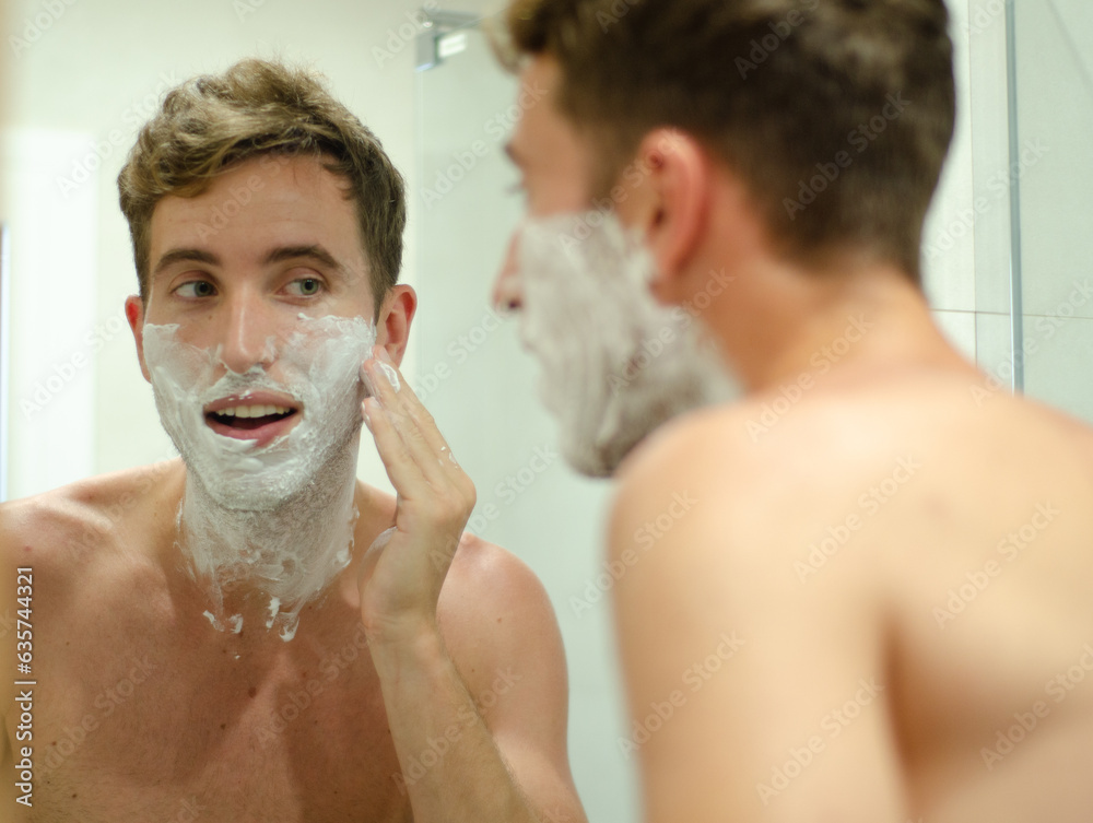 Young man smiling and looking at himself in the mirror while shaving.