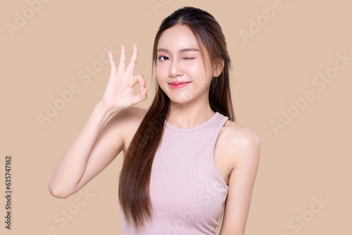 Beautiful young Asian woman with healthy and perfect skin shows OK signal on isolated brown background. Facial and skin care concept for commercial advertising.