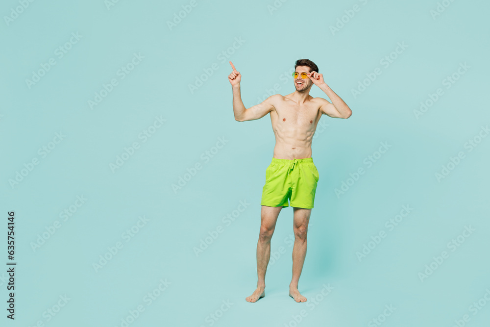 Obraz premium Full body young man wear green shorts swimsuit sunglasses relax near hotel pool look aside on area mockup point finger aside isolated on plain blue background Summer vacation sea rest sun tan concept