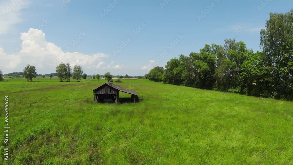 Overflight of a green field with blue sky and white clouds. There are trees in the background and a small wooden hut