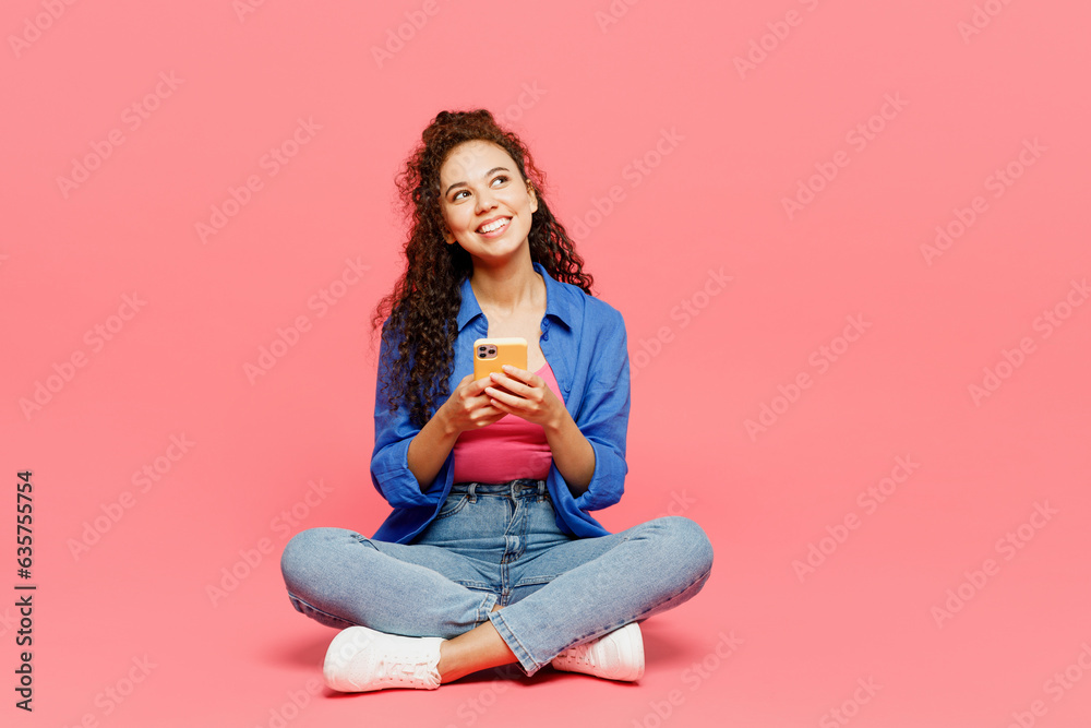 Full body young woman of African American ethnicity wear blue shirt casual clothes sit use mobile cell phone look overhead on area isolated on plain pastel pink background studio. Lifestyle concept. Full body young woman of African American ethnicity wear blue shirt casual clothes sit use mobile cell phone look overhead on area isolated on plain pastel pink background studio. Lifestyle concept.