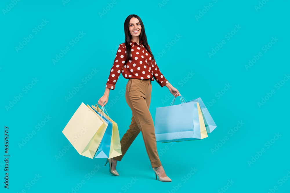 Full size portrait of stunning positive girl wear elegant clothes hold store bags walking isolated on turquoise color background Full size portrait of stunning positive girl wear elegant clothes hold store bags walking isolated on turquoise color background