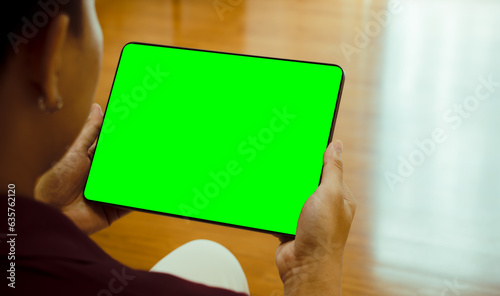 Mockup image of asian man holding black digital tablet with blank green screen at home or office.