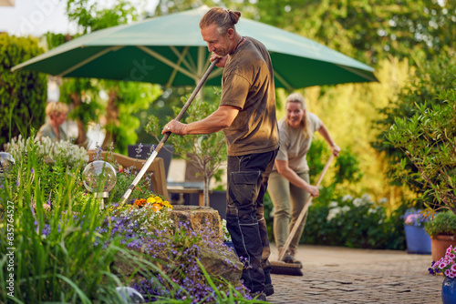 Male gardener raking the the flower bed while the collegue is sweeping the terrace in summer