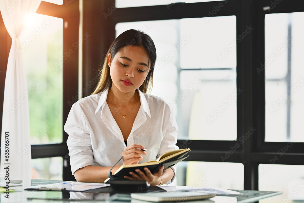 Obraz premium Millennial female employee checking work schedule plan on daily planner while sitting at modern workplace.