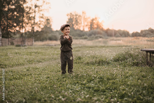 A boy is playing on a farm or ranch field, resting. Portrait of a little boy in a cap and overalls. Childhood. An authentic picture with candid emotions. © Anastasia