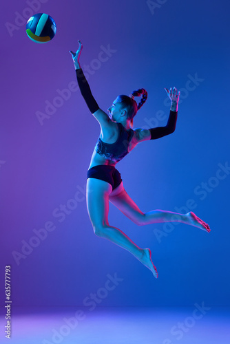 Dynamic full-length image of professional female athlete, woman playing volleyball against blue studio background in neon light. Concept of professional sport, competition, health, hobby, action, ad