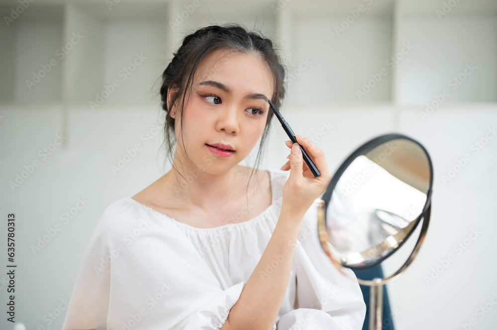A beautiful Asian female looks in the mirror and draws her eyebrows with a brow pencil A beautiful Asian female looks in the mirror and draws her eyebrows with a brow pencil