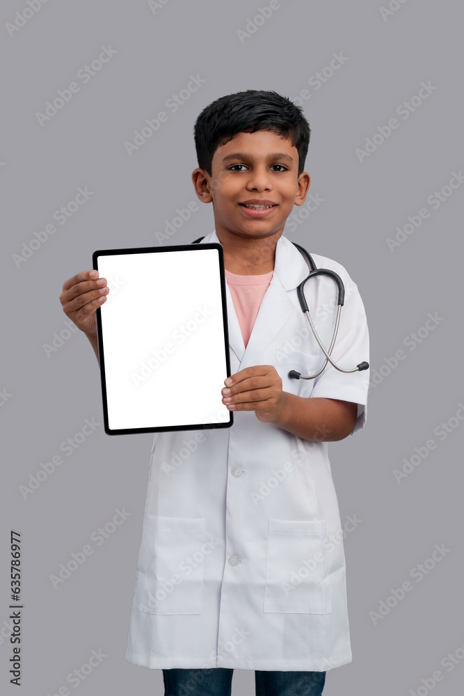 Indian Asian kid boy aged 7 to 8, wearing a doctor apron with stethoscope. Holding tablet or Ipad in hand. He had a dream to future study as Doctor. He smiled happily, Concept little Doctor