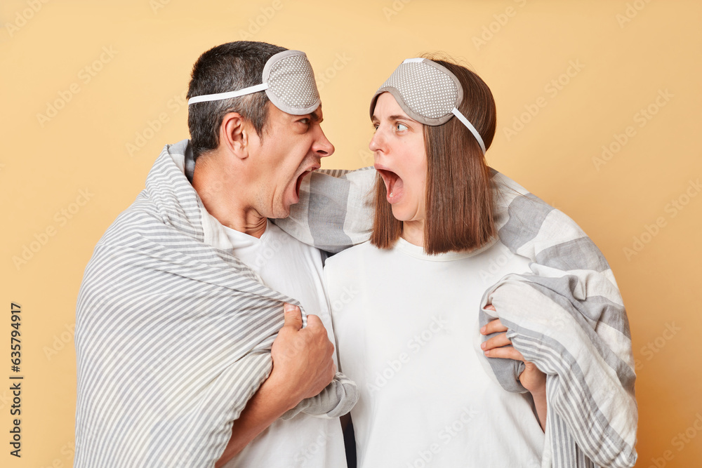 Shocked crazy couple man and woman wrapped in blanket isolated over beige background being oversleep screaming with shocked expression.