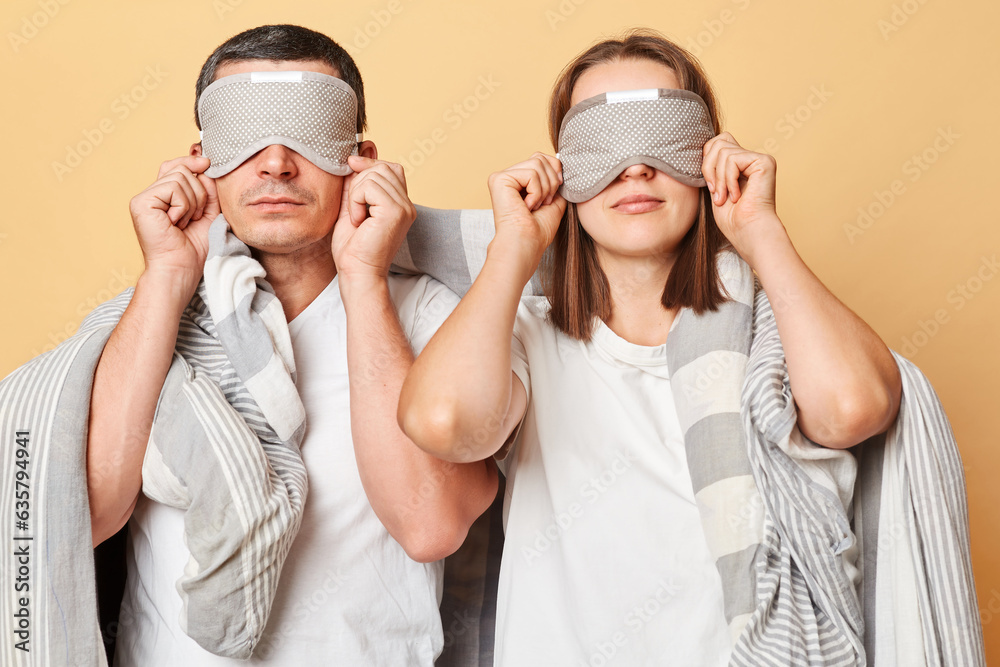 Serious tired exhausted couple man and woman wrapped in blanket isolated over beige background covering eyes with blindfold have lack of sleep Serious tired exhausted couple man and woman wrapped in blanket isolated over beige background covering eyes with blindfold have lack of sleep