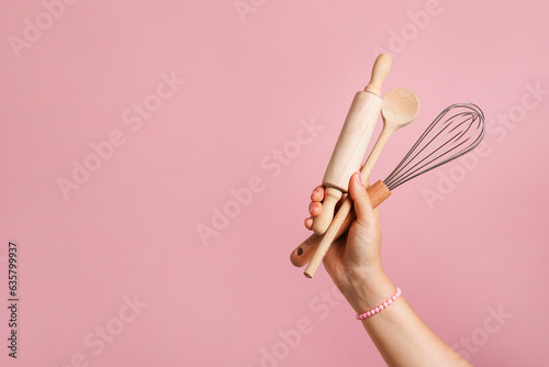 Female hand holding kitchen utensils for food and bakery on pink background. whisk, rolling pin and wooden spoon