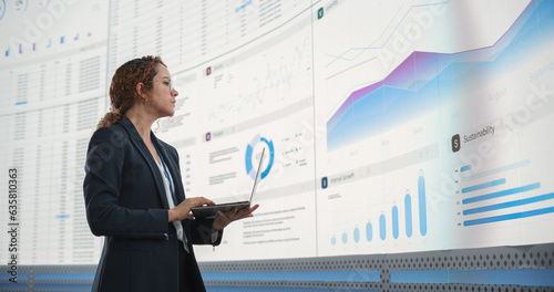 In the Monitoring Office: Successful Hispanic Businesswoman Holding Laptop Computer And Analyzing Data On Big Digital Screen With Graphs and Charts. Female IT Entrepreneur Running Software Company. photo