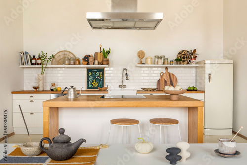 Warm and cozy kitchen interior with mock up poster frame, wooden kitchen island, barstool, fridge, white kitchen furniture, gray pitcher, salt cellar and kitchen accessories. Home decor. Template.