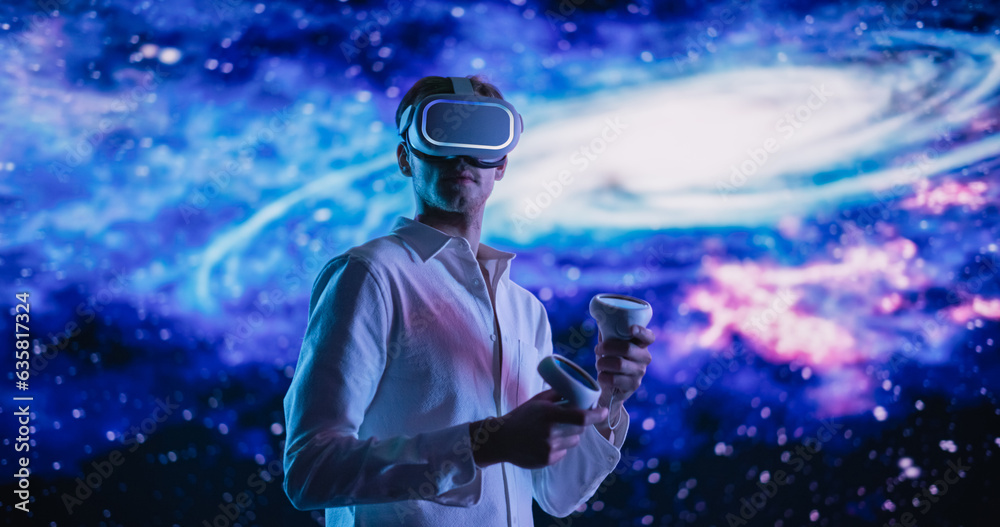Young Male Wearing a Virtual Reality Headset and Using Controllers in a Futuristic Room with Digital Space Animation. Scientist Exploring VR Universe, Different Galaxies, Planets, Space and Time