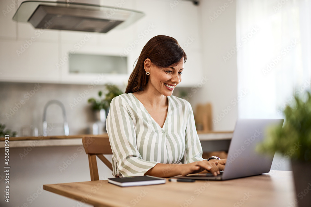A smiling Caucasian woman typing on a laptop at the kitchen table at home. A smiling Caucasian woman typing on a laptop at the kitchen table at home.