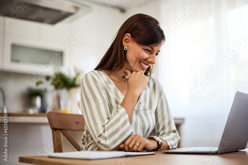 Beautiful smiling woman looking at laptop while sitting at wooden table in the bright kitchen.