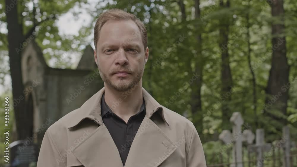 Medium portrait of serious young Caucasian man wearing long trench coat standing at cemetery at daytime and looking at camera Medium portrait of serious young Caucasian man wearing long trench coat standing at cemetery at daytime and looking at camera