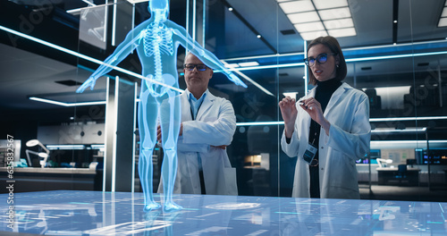 Two Bioengineers Working With Computer-Powered VFX Hologram Of Human Body In Futuristic Lab. Man And Woman Researching Bone Structure, Developing Innovative Technological Healthcare Solutions.