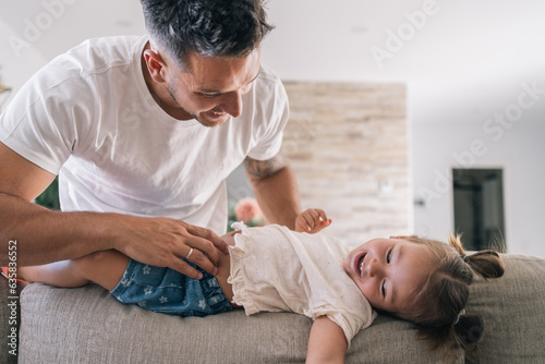 Young smiling father tickling his happy little daughter with two lock of hair on the back of his living room sofa. photo