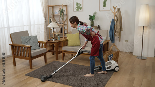 full length young asian chinese woman having back ache from cleaning apartment. illness wife vacuuming floor in living room. tired female householder using vacuum cleaner on carpet.