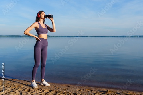 A young woman in a tracksuit drinks water after a running workout on the beach. © makedonski2015