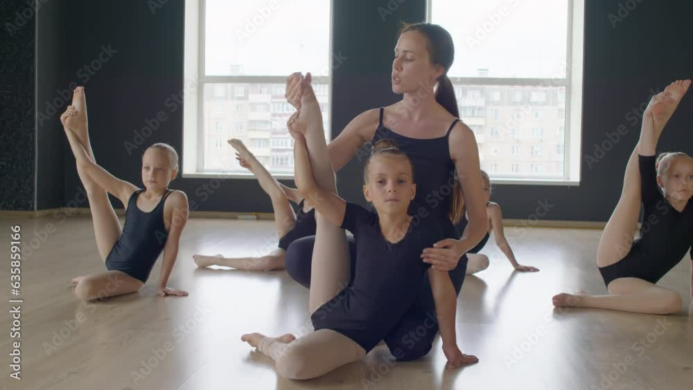 Young coach helping flexible girl to do gymnastic exercise with raised leg while sitting on floor in studioYoung coach helping flexible girl to do gymnastic exercise with raised leg while sitting on f