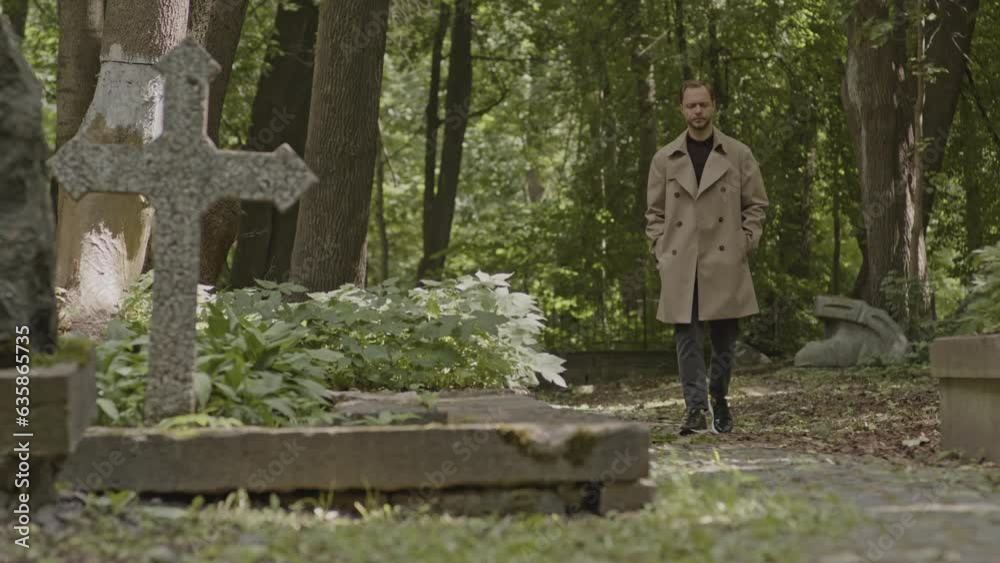 Young Caucasian man in camel trench coat strolling along cemetery at daytime Young Caucasian man in camel trench coat strolling along cemetery at daytime