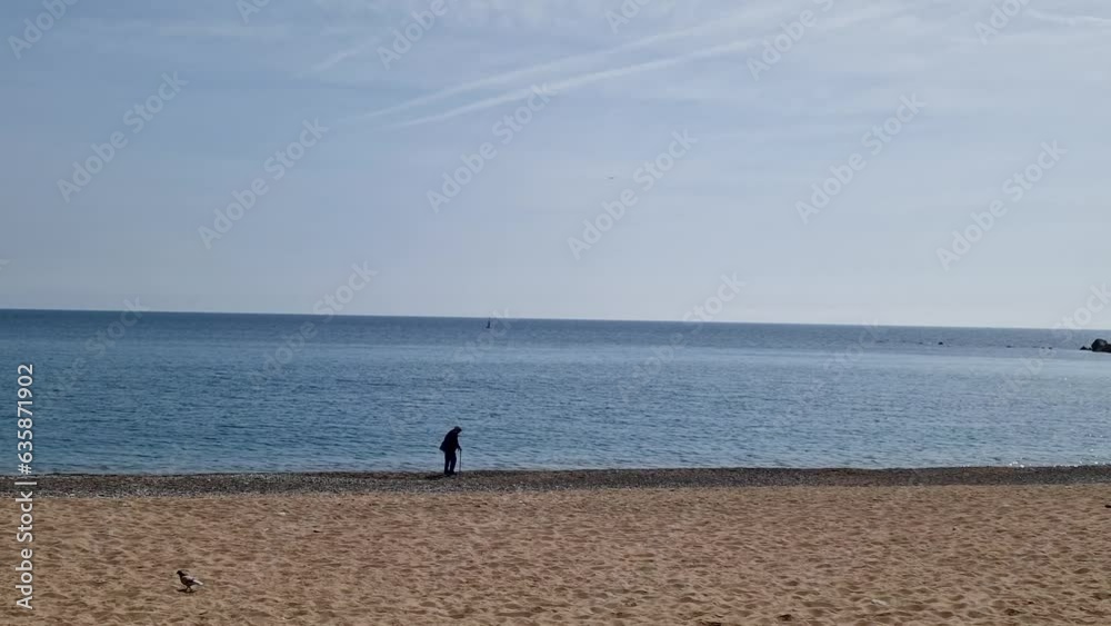 An elderly man with a stick walks along the beach in Barcelona. Pensioner's walk on the yellow sand of the beach by the blue sea An elderly man with a stick walks along the beach in Barcelona. Pensioner's walk on the yellow sand of the beach by the blue sea