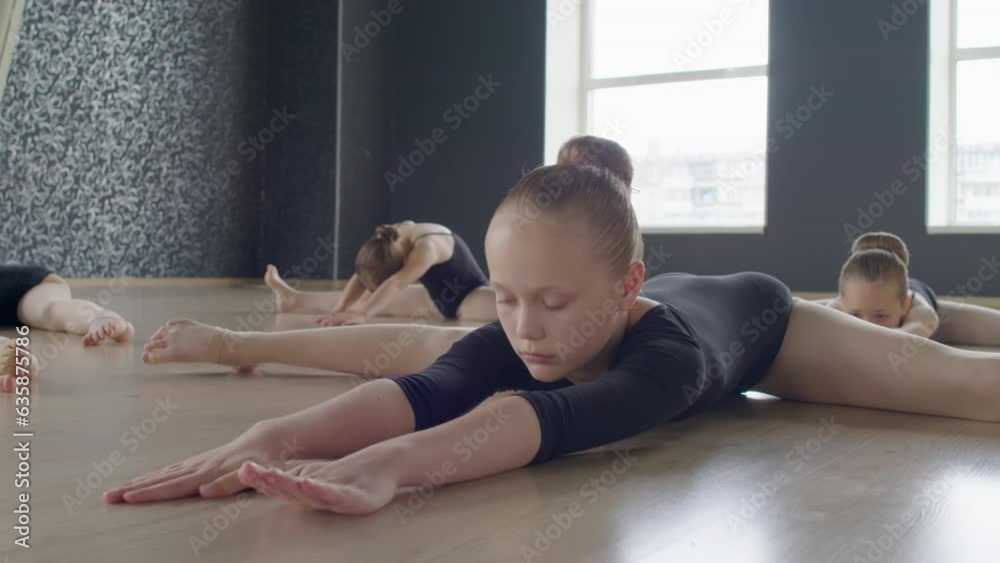Medium long shot of little ballerinas in leotards doing stretching exercises on floor in studio