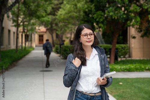 smiling asian korean college girl walking on leafy school campus in California usa. she is carrying a textbook while going to class