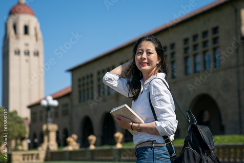 attractive asian Chinese female exchange student holding book and pushing hair back while looking at camera with a smiling face at school university with hoover tower at background photo