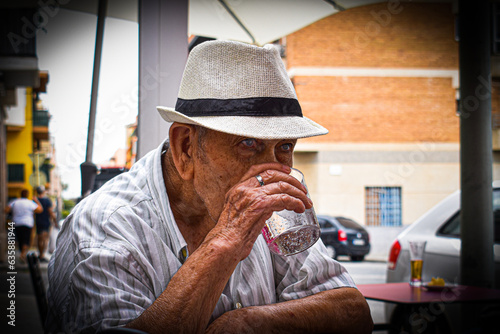 Abuelo anciano hombre mayor con gorro, tomando un refresco o un vaso de agua sentado en una terraza