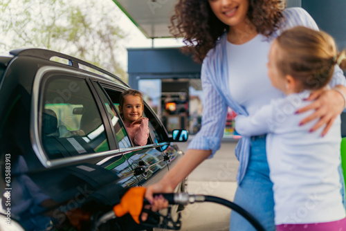 A mother pours fuel into the car while her daughter hugs her and a younger one looks at them, both are happy